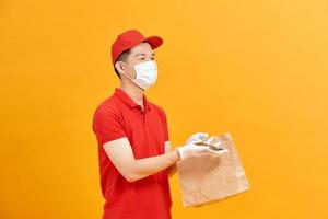 repartidor sosteniendo una bolsa de papel con comida sobre fondo blanco, repartidor de comida con máscara protectora y guantes protectores foto