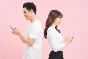Romantic messages. Beautiful young loving couple holding mobile phones and standing back to back against pink background photo