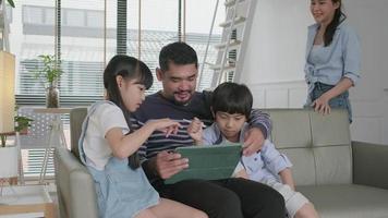feliz familia tailandesa asiática, padres e hijos se divierten usando una tableta digital juntos en el sofá de la sala de estar, un hermoso fin de semana de ocio y un estilo de vida de bienestar doméstico con tecnología de Internet.