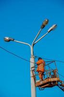 A municipal worker in protective equipment performs hazardous work to eliminate an interruption in the power grid. A worker repairs a street lamp from an aerial platform basket. photo