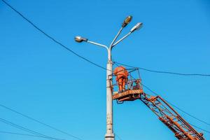 A municipal worker in protective equipment performs hazardous work to eliminate an interruption in the power grid. A worker repairs a street lamp from an aerial platform basket. photo