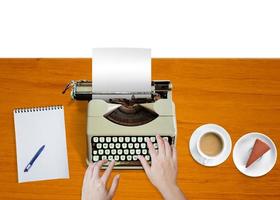 Woman's hand typing on an old typewriter, notebook, coffee mug, plate of cakes on a wooden table, top view. photo