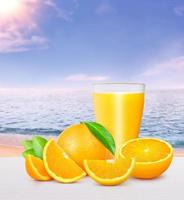 a glass of orange juice oranges and oranges cut into pieces with green leaves On a white wooden floor there is a beautiful sea and sky in the background. photo