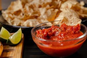 cierre la mano con salsa de tomate con chip dip con plato con taco, chips de nachos y salsa de tomate. nachos picantes sobre fondo de madera rústica foto