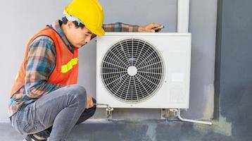 el técnico de aire acondicionado usa una llave para apretar la tuerca del compresor de aire. un joven reparador asiático revisando una unidad de aire acondicionado exterior. foto