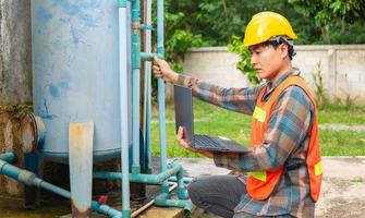 Engineer working in drinking water factory using a laptop computer to check water management system and boiler water pipe  outside the factory photo