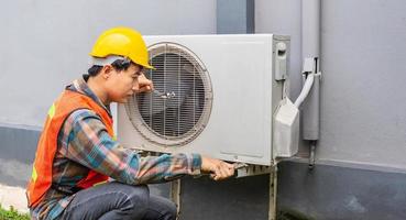 el técnico de aire acondicionado usa una llave para apretar la tuerca del compresor de aire. un joven reparador asiático revisando una unidad de aire acondicionado exterior. foto