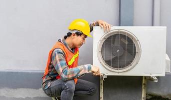 el técnico de aire acondicionado usa una llave para apretar la tuerca del compresor de aire. un joven reparador asiático revisando una unidad de aire acondicionado exterior. foto