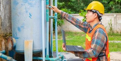 Engineer working in drinking water factory using a laptop computer to check water management system and boiler water pipe  outside the factory photo