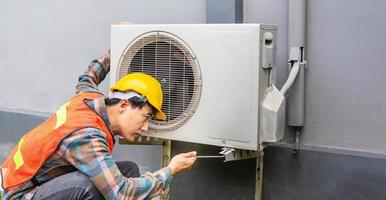 el técnico de aire acondicionado usa una llave para apretar la tuerca del compresor de aire. un joven reparador asiático revisando una unidad de aire acondicionado exterior. foto