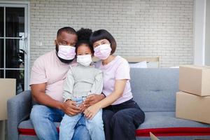 African American family moves into a new home, sitting on the sofa in the living room. They wear dust masks and protect against coronavirus. Concept of mixed race family. Copy space photo