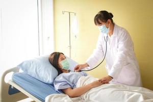 Female doctor holding a stethoscope, doctor examining the heart, Asian woman patient Wear a blue mask and lie in a hospital bed. Concept of medical service, health insurance. Copy space photo