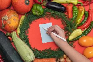 las verduras se disponen alrededor de una hoja de papel y un lápiz. espacio vacío para texto. mano femenina escribiendo una receta en un espacio en blanco vacío sobre un fondo rojo. foto