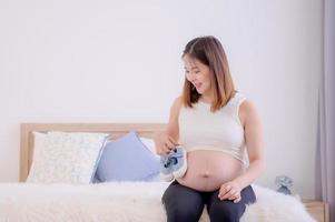 A pregnant Asian woman sits on the bed and provides a small pair of shoes for her child photo