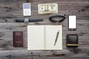 cosas de viaje para viajar. Pasaporte, billetera, dinero, teléfono, gafas de sol, reloj, lista de tareas en una mesa de madera. cuaderno abierto con página en blanco. foto