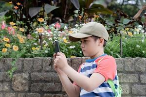 niño de escuela usando teléfono móvil tomando fotos de flores en el jardín, niño aprendiendo sobre la naturaleza en el parque, niño feliz divirtiéndose disfrutando de actividades al aire libre en un cálido día de verano