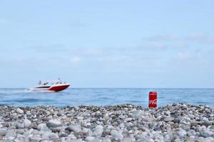 ANTALYA, TURKEY - MAY 18, 2021 Original Coca Cola red tin can lies on small round pebble stones close to sea shore. Coca-cola can and speed boat on beach photo