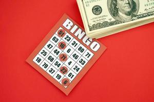 tablero de bingo rojo o naipe para ganar fichas y pila de billetes de dólar. Tarjeta de bingo clásica americana o canadiense de cinco a cinco sobre fondo rojo. foto