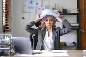 Portrait of beautiful asian woman at the desk with paperwork on her head. business overwhelmed stressed concept. photo