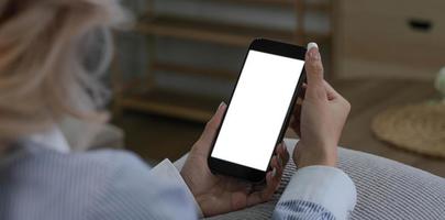 imagen de primer plano, hermosa joven asiática usando su smartphone mientras se relaja en su sala de estar mínima. una mujer sosteniendo una maqueta de pantalla blanca de teléfono móvil. foto