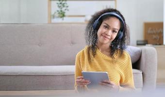 Young satisfied female in casual clothes yellow and headphones surfing digital tablet while sitting on floor with legs crossed near soft couch in light modern living room photo