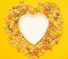 empty white plate in the shape of a heart on a background of colored pasta. restaurant menu concept. festive serving of dishes. yellow background. Design template. Place the symbol photo