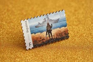 TERNOPIL, UKRAINE - SEPTEMBER 2, 2022 Famous Ukrainian postmark with russian warship and ukrainian soldier as wooden souvenir on golden glitter background photo