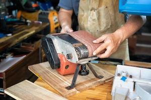 carpintero que usa delantal e instala o quita la correa abrasiva de la lijadora de banda en tabel de madera en el taller, el fabricante de bricolaje y el concepto de carpintería. enfoque selectivo foto