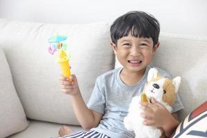 Adorable happy Asian little boy looking at the camera wearing a gray shirt and blue-white striped shorts sitting on a cream sofa is smiling happily. Left hand dog doll and right hand holds a toy photo