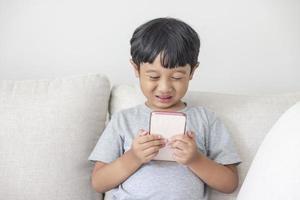 An adorable happy Asian boy wearing a gray shirt and blue-white striped shorts is having fun playing with his smartphone on a cream sofa. looking at the mobile screen photo