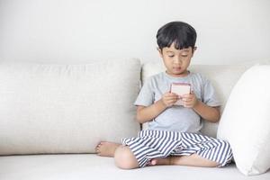 An adorable happy Asian boy wearing a gray shirt and blue-white striped shorts is having fun playing with his smartphone on a cream sofa. looking at the mobile screen photo