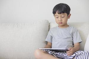 adorable niño asiático feliz con una camisa gris y pantalones cortos de rayas azules y blancas se divierte jugando con su tableta en un sofá color crema. mirando la pantalla del movil foto