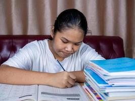 girl doing homework on a wooden table and there was a pile of books next to it The background is a red sofa and cream curtains. photo