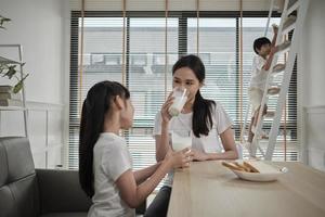 A Healthy Asian Thai family, little daughter, and young mother drink fresh white milk in glass and bread joy together at a dining table in morning, wellness nutrition home breakfast meal lifestyle. photo