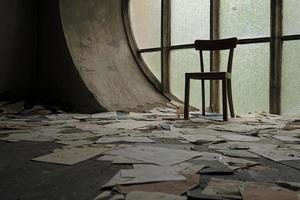 Chair in front of a round window in an abandoned church with papers and book pages lying on the ground photo