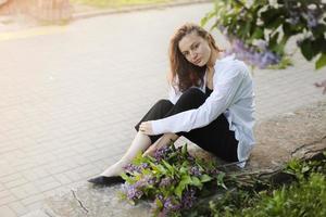 young beautiful girl in a white shirt and black pant sitting near blooming lilac trees with purple flowers outdoors, flowering tree, spring, sun, happiness, tenderness. photo