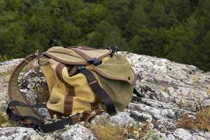 A brown-green backpack lies on a large gray stone on a rock against the backdrop of green mountains. photo