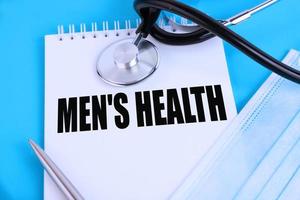 Men's Health, text written in a notebook lying on a blue background, with a stethoscope and a medical mask. Medical concept.