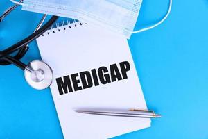 MEDIGAP, text written in a notebook lying on a blue background, with a stethoscope and a medical mask. Medical concept. photo