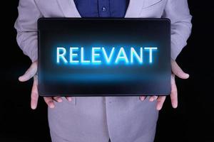 RELEVANT text, word written in neon letters on a black laptop, in front of a businessman in a gray suit. photo