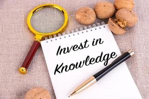 Invest in knowledge, the text is written in a notebook, next to a pen, a magnifying glass and walnuts on a linen background. photo