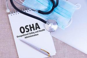 OSHA Occupational Safety and Health Administration, the text is written in a notebook, next to a pen, a disposable medical mask and a laptop on a linen background. photo