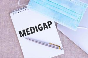 MEDIGAP, the text is written in a notebook, next to a pen, a disposable medical mask and a laptop on a linen background. photo