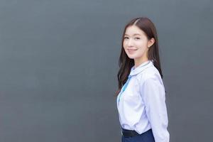 hermosa estudiante asiática de secundaria con uniforme escolar con sonrisas confiadas mientras mira a la cámara felizmente con gris en el fondo. foto