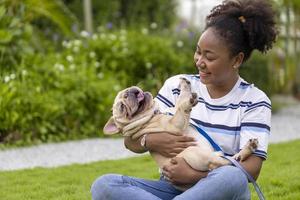 African American woman is playing with her french bulldog puppy while walking in the dog park at grass lawn after having morning exercise during the summer photo