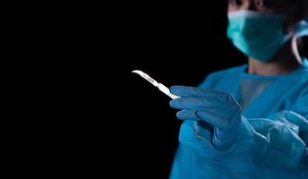 Hand of surgeon in blue medical glove holding a scalpel, express emotion feeling stress before operation. Tools medical instrument. Black background underexposure for shadow environment copy space photo