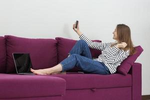 young woman uses social apps and takes photo herself in cellphone in home. Woman working, learns and using laptop computer in the bedroom. Freelance. Communication and technology concept.