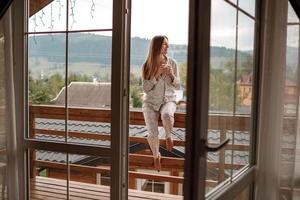 Young woman on the balcony holding a cup of coffee ore tea in the morning. She in hotel room looking at the nature in sumer. Girl is dressed in stylish nightwear. Relax time. photo