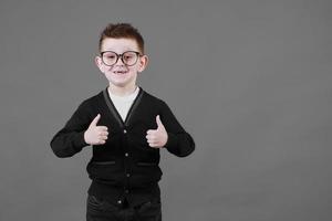 Like. Portrait of happy little schoolboy with glasses smiling at camera and doing thumbs up gesture, showing agree cool approval sign. indoor studio shot isolated on gray background photo