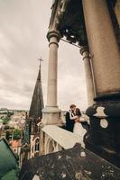 Happy newlywed. beautiful bride and stylish groom are kissing on the balcony of old gothic cathedral with panoramic city views photo